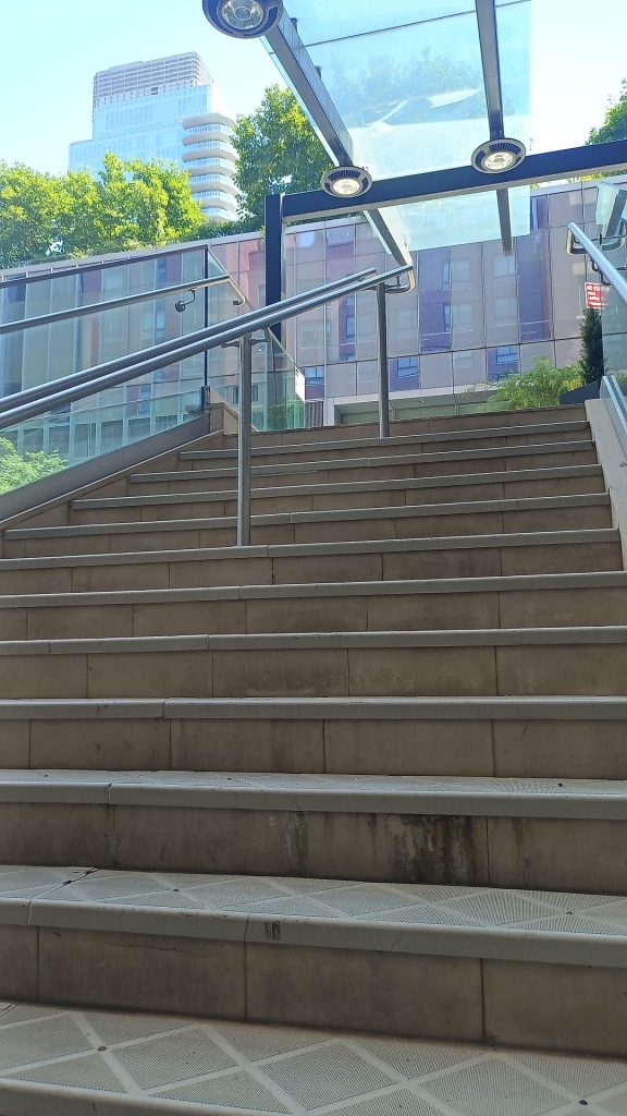 Das Foto zeigt eine breite Außentreppe, die nach oben zu einem Ausgang oder Übergang führt. Die Treppe besteht aus hellbraunen Stufen mit einem rutschfesten Belag an den Vorderkanten. In der Mitte und an beiden Seiten der Treppe sind silberne Handläufe aus Metall angebracht. Über der Treppe befindet sich ein Glasdach mit integrierten runden Lampen, das von einem schwarzen Metallrahmen gehalten wird.
Im Hintergrund sind moderne Gebäude mit Glasfassaden zu sehen, hinter denen teilweise grüne Bäume hervorschauen. Über den Bäumen ragt ein hohes, modernes Hochhaus mit runden Balkonen in den Himmel. Die Szene ist bei Tageslicht aufgenommen, der Himmel ist klar und blau. Sonnenlicht scheint auf die oberen Teile der Bäume und Gebäude.