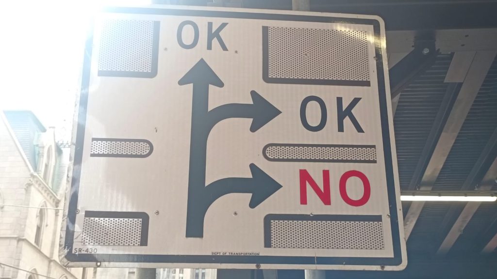 Das Foto zeigt ein großes Verkehrsschild aus Metall, das unter einem Überdach hängt. Auf dem Schild sind drei dicke schwarze Pfeile abgebildet, die unterschiedliche Fahrtrichtungen zeigen: Einer geht nach links oben, einer geradeaus und einer nach rechts. Über dem linken und dem mittleren Pfeil steht jeweils in großen Buchstaben „OK“. Über dem rechten Pfeil, der eine abwärts gebogene Rechtskurve anzeigt, steht hingegen „NO“ in roter Schrift. Der Hintergrund ist weiß, die Pfeile und der Rahmen dunkelblau. Rund um das Schild sind reflektierende, silberne Flächen eingelassen. Im unteren Rand steht klein: „DEPT OF TRANSPORTATION“. Im Bildhintergrund ist ein historisches Steingebäude mit Spitzdach zu sehen.